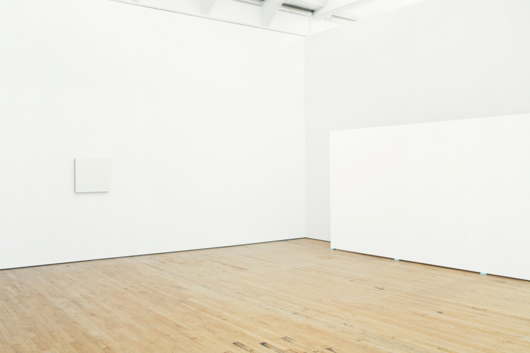 A small, square white painting hangs on a wall above a wood floor. A partially visible, large, rectangular white painting rests on blue pads on the floor near the wall to the right.