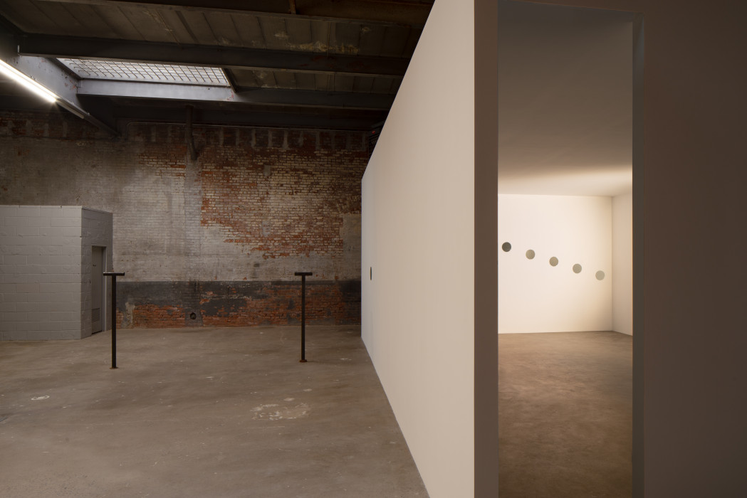 An industrial space bisected by a white wall, with two metal sculptures in the room on the left, each a few feet tall and shaped like the letter 'T', and five holes in the back wall of the room on the right, descending to the right.