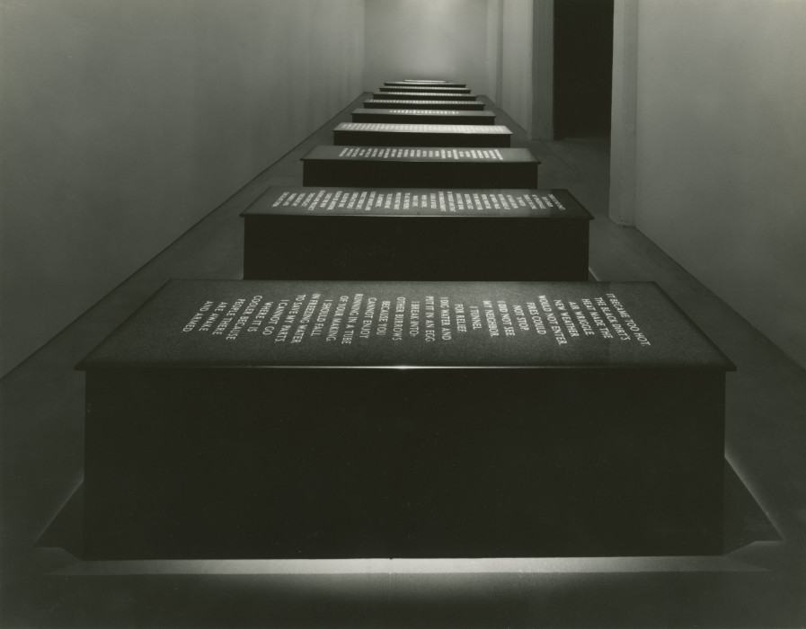 Ten black, rectangular marble platforms with all caps text carved into the top face of the platform sit in a single line in a narrow room. Each platform has a light shining on its face illuminating the text.
