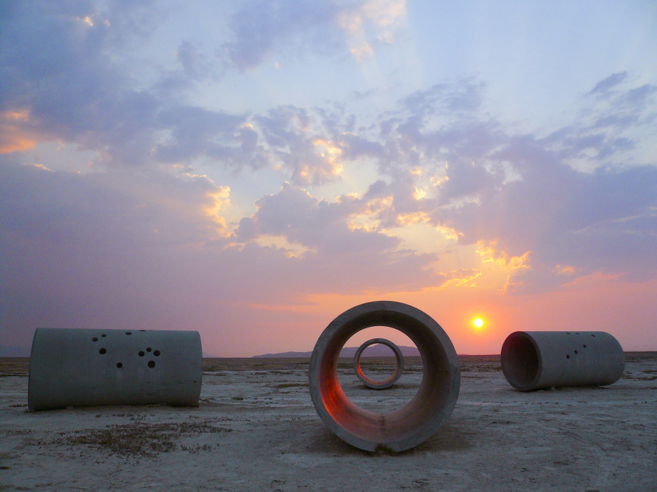 Four large, concrete tubes sit in a spread out X-formation on a sprawling desert basin ground with a mountain range in the background. One tube looking directly into another tube and the other two tubes sit in profile. Each tube has various sized circles drilled through the concrete in a constellation pattern. The sun is beginning to set against a pastel red, pink, blue, and purple sky with a spread of clouds.