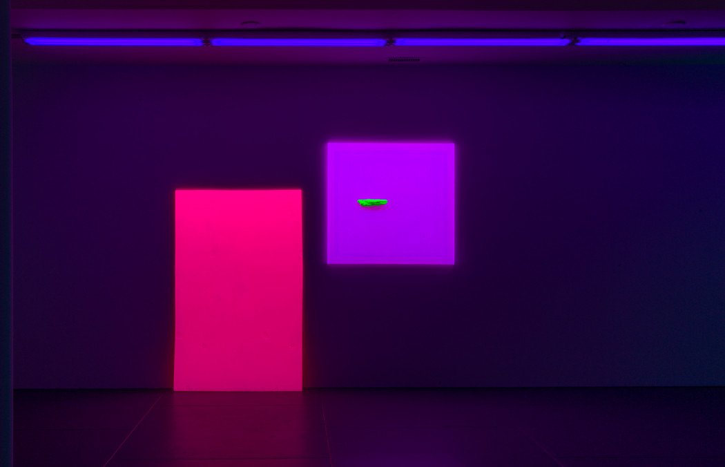 A vertical fluorescent magenta rectangle leans against a wall to the bottom left of a fluorescent purple square painting with a small, neon green piece of driftwood floating off-center. The room is dark and illuminated by a line of violet florescent lights on the ceiling.