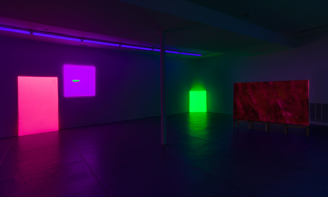 Four fluorescent geometric forms occupy a dark room. On the back wall there is a vertical magenta rectangle leaned against it and a purple square painting with a small, neon green piece of draftwood floating off-center. In the back corner there is a vertical green rectangle leaned against it. All three forms are under a line of violet fluorescent lights. To the right is a large horizontal magenta rectangle with dark brushstrokes on wooden stilts.