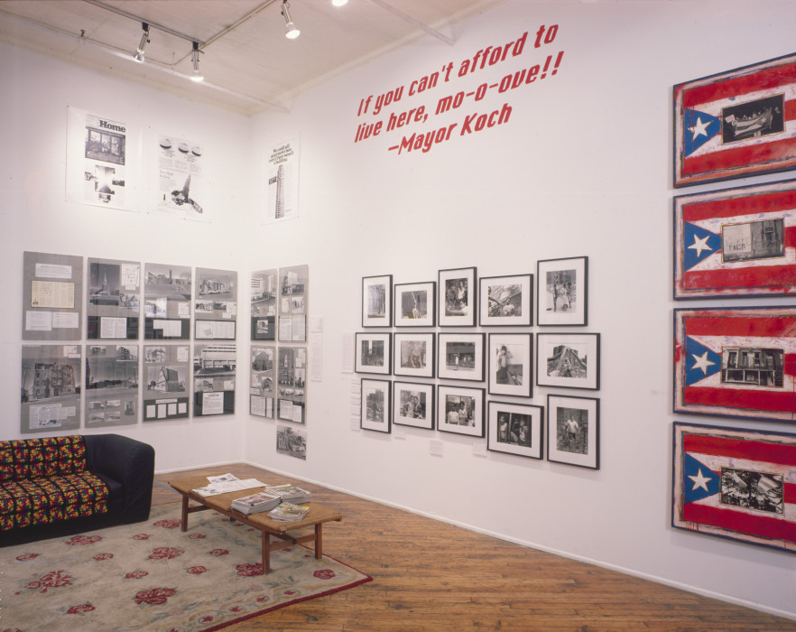 An installation view of several groupings of works hung on two walls, including charts, collages, black and white photographs, and four paintings of the Porto Rican flag with a different photo in the center of each. In the center of the room, there is a couch atop a rug with a small coffee table with magazine nearby. On one wall, there is a red vinyl text that reads 