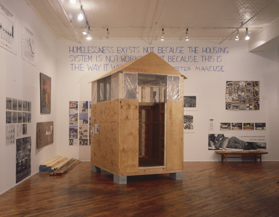 A small house-like structure made of plywood and plastic in the center of a room. The walls of the room have various collages, photographs, drawings on cardboard, and charts hung on the walls, as well as the following text, partially obscured by the structure: 
