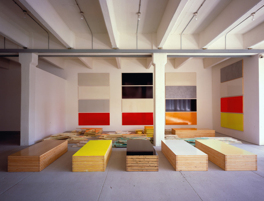 Evenly stacked piles of rectangular strips of wood painted different colors sit on a floor, while other painted rectangles hang horizontally on the walls.