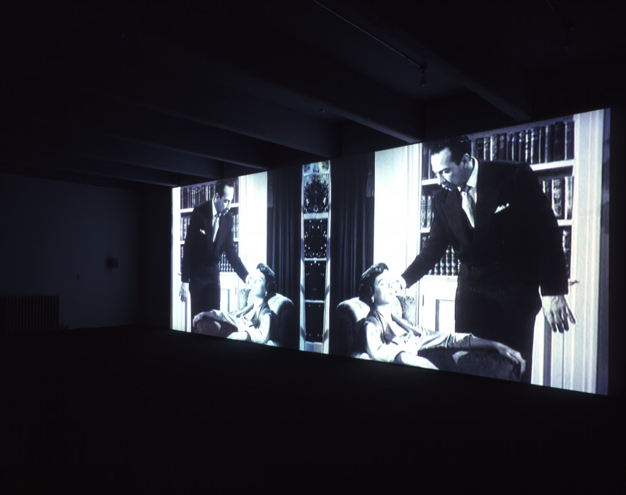 Two identical black-and-white images of a woman seated in a chair and a man leaning towards her are projected in a dark space.