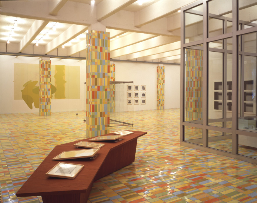 Multicolored mosaiced floor and columns in a gallery space with artworks on the walls and on a benchlike structure in the foreground.