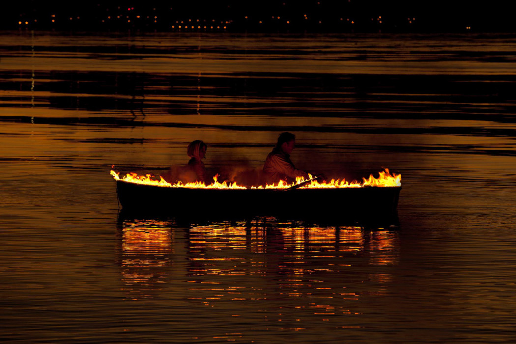 A burning boat on a body of water, reflecting black and orange, with two people sitting in it, one rowing.