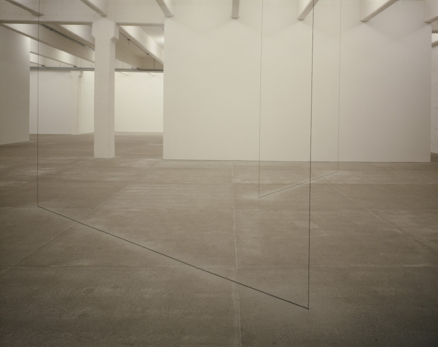 A thin black line stretches diagonally across a cement floor and connects to two other thin black lines that stretch toward the ceiling in a white room. Another similar shape is placed a few feet away and extends perpendicularly into the background.