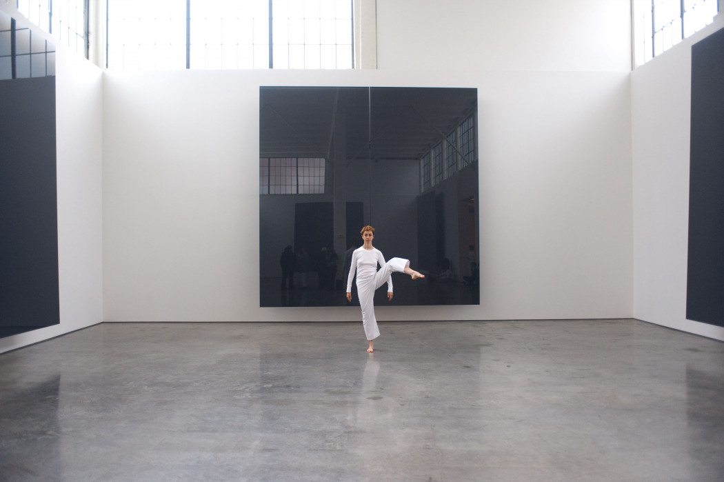 A dancer dressed in white, facing forward with their right leg raised and extended to the right in a curve, standing in front of a large and mirrored black square work installed on a white wall.