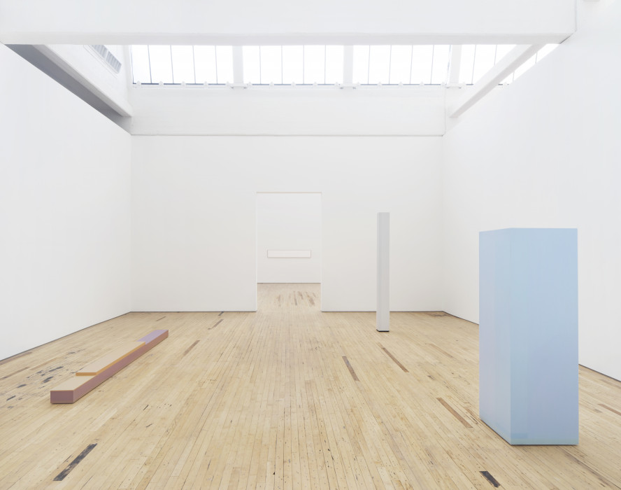 A room with three sculptures: a low and flat rectangular tan and lilac work on the left, a narrow off-white rectangular pillar on the right in the back, and a wide and light blue pillar in the right foreground. There is a white rectangular painting visible through a doorway in the back.