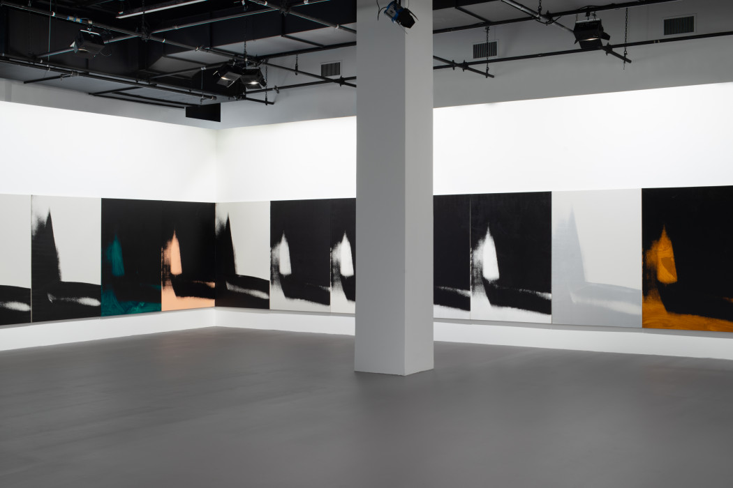 The corner of a gallery with a gray pillar in the center, with both walls lined with many iterations of the same vertically oriented print of an abstracted and vague shadow form that descends on the left and moves across the bottom edge. Each print is in black and one other color, either oragne, dark green, peach, light grey, or white, except for one on the left with a light grey background and a slightly darker grey shadow.