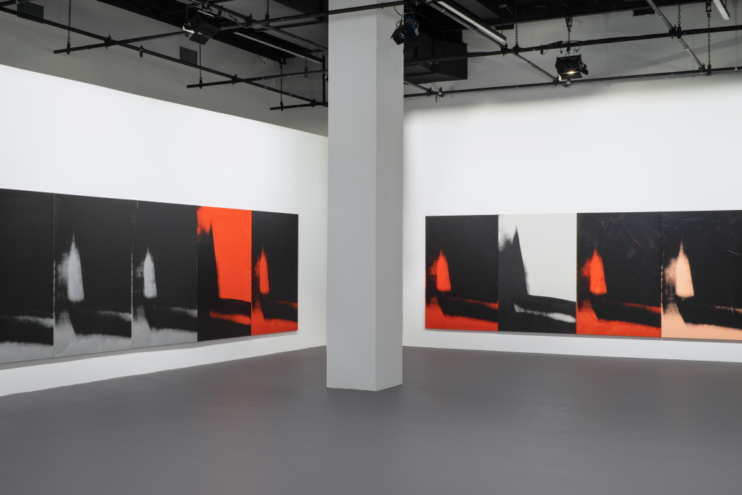 The corner of a gallery with a gray pillar in the center, with both walls lined with many iterations of the same vertically oriented print of an abstracted and vague shadow form that descends on the left and moves across the bottom edge. Each print is in black and one other color, either grey, red or peach.