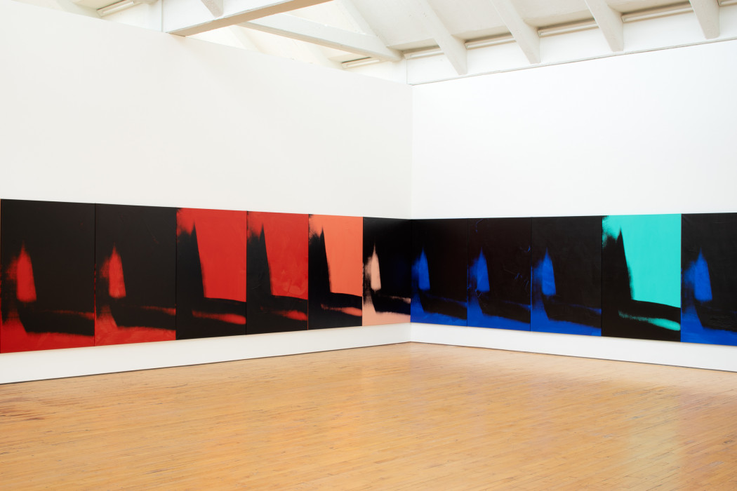 The corner of a gallery, with both walls lined with many iterations of the same vertically oriented print of an abstracted and vague shadow form that descends on the left and moves across the bottom edge. Each print is in black and one other color, either red, blue, peach, orange, or aqua.