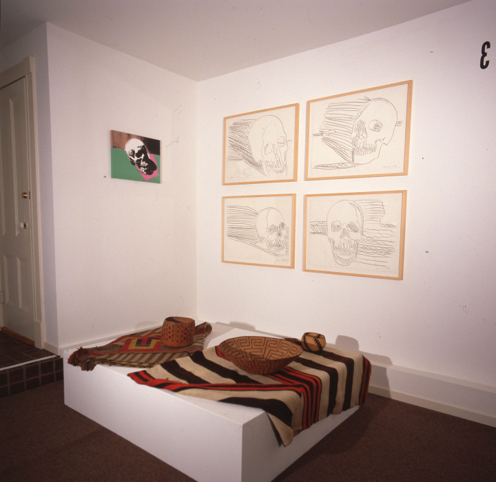 A corner of a gallery is shown with a small colorful painting of a skull on the left wall, 4 framed pencil drawing in a grid on the right wall, and a platform positioned in front with blankets and woven baskets on top.