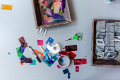 Two pairs a scissors, a box of film slides, a marker, tape, and transparently colored plastic swatches litter a table.