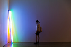 A neon sculpture leans in a corner and glows blue, green, orange, and white. A person views the light from afar.