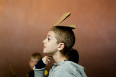 A boy in profile points his right hand to his neck and balances a brown paper folded in half on top of his head in front of peers and a caramel background.
