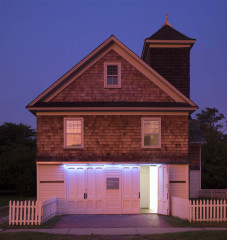 A small building with two sets of double doors and a white picket fence, one door is open revealing a lit entryway inside. There are two windows on the floor above, a 3rd window in the attic, and a structure like a steeple on the back right side. The sky is a purple blue.