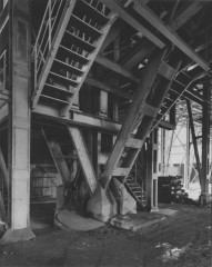 Black-and-white photograph, taken at an angle, of the underside of a staircase and other diagonal building supports.