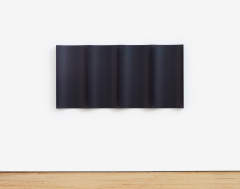 A black metal rectangular sculpture mounted on a white wall. The sculpture is a black metal sheet that is bent and folded into a symmetrical zig zag, installed so that the form alternates between extending into the gallery and then receding to touch the wall, starting and ending off of the wall.