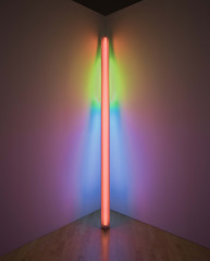 A long, orange, fluorescent lightbulb leans against an illuminated corner of a room that appears green, blue, and pink.