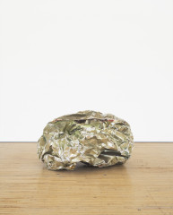 A crumpled ball-like sculpture made from reflective silvery green metal parts rests on a wooden floor.
