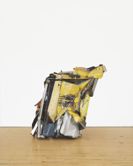 A box-shaped sculpture made of yellow, blue, and silver metal parts rests on a wooden floor.