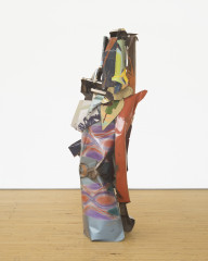 A vertically oriented sculpture made of multicolored metal parts rests on a wooden floor.