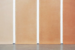 Four long paper works installed in a row on a white wall, the bottoms dragging slightly on the concrete floor. They are all in tan and orange shades, arranged from lightest to darkest staring on the left. The top edges and the two drawings on either end are slightly truncated.