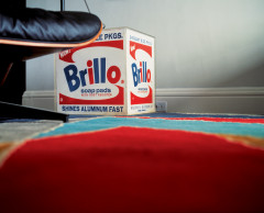 Photograph of a Brillo box and the lower part of a chair on a multicolored carpet.