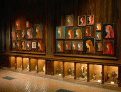 Many different reproduction paintings of a left-facing side profile portrait of person wearing a red headscarf hang on a wooden wall. Below the paintings on the ground are inset wall vitrines displaying figurines and decorative plates.