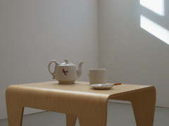 A white china teapot with a small image of bird on it, a white mug, and a white sauce holding a lit cigarette, all sitting atop a small curved wooden table.