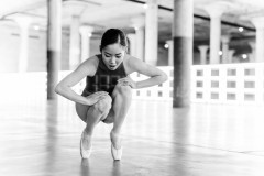 Black-and-white photograph of dancer hunched over while squatting en pointe and backlit with a neon, gridded light installation running the length of the floor in the background.