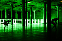 Two dancers on opposite sides of the floor are hunched over while crouched en pointe, backlit with a neon green, gridded light installation running the length of the floor in the background..