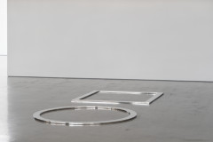 A flat outline of a circle next to a flat outlone of a square, both in shiny stainless steel, rest on a cement floor with a white wall in the background.
