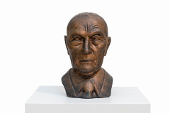 A bronze bust of an older bald man wearing a suit and tie, facing forward and displayed atop a white pedestal.