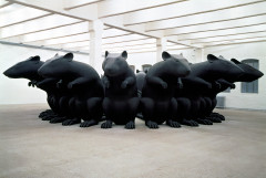 A ring of very large black rat figures, arranged around a pillar in a white room, all facing outwards.