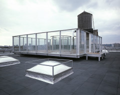 A roofless glass room on a platform installed on a rooftop, with a water tower just behind it, and the tops of square skylights surrounding it.