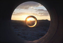 A large, concrete tube oculus looks out onto another concrete tube directly across from it on a desert basin. The background is the sun setting against a mountain range.
