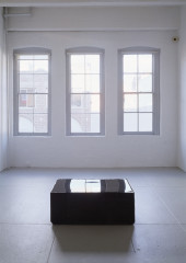 A large, glossy black rectangular prism sits in the center of an empty white room facing three windows that let in natural daylight.