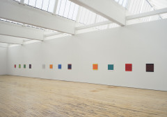 Twelve small square paintings installed in a row on a white wall, grouped in three groups of four. Each unique painting is of one solid color with the top and bottom edges in a second contrasting color.