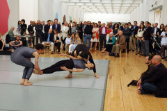 A dance performance with three dancers on a gray mat is observed by a crowd of people in a gallery space.