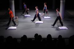 A group of five dancers on stage in black trousers and different colored and patterned tops step in identical motion towards stage right, sandwiched between a metal folding chair behind them and a white pillow sitting in front of them.