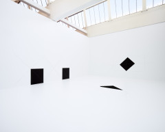 A room is entirely painted white, including the floor, and two black squares, one diamond, and one triangular have been painted in different parts of the room along with light geometric line drawings.