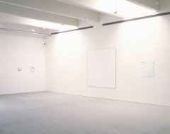 Two small and predominately white paintings appear in the corner of a white room. A barely discernible white square, a large white painting, and another white square hang in a row on the other wall.