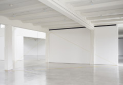 Three thin yellow lines form the outline of a triangle that consumes most of the space of a white room with a concrete floor. Two thin black lines form a right angle in an adjacent room.