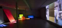 Multicolored, differently lit geometric forms sit in an unlit room to one side while a geometric grid is projected onto the opposite wall.