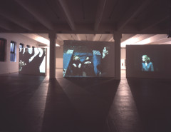 Images of people are projected onto three different large panels in a empty, white space.