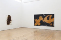 Two works installed on perpendicular white walls; the one on the left is a white vertical rectangle with a large dark brown shape descending in the center, with a horizontal shelf across the center of the work, and the work on the right is a long rectangular painting with a black background and warm brown abstract shape across it.